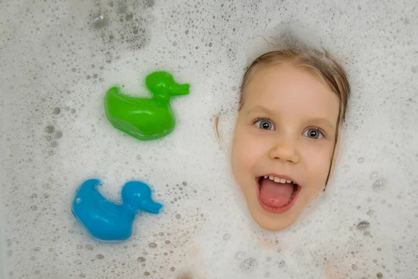 Das fröhliche Gesicht eines kleinen Kindes im Schaumstoff der Badewanne neben den Spielzeugenten. Blick von oben. Kinderhygiene. Badespaß. — Stockfoto