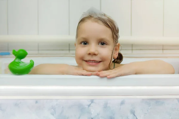Gyermekhigiénia. Egy gyönyörű kislány, habbal a fején, és egy zöld kacsa fürdésre néz ki a kádból. A gyermek édesen fürdik és mosolyog.. — Stock Fotó