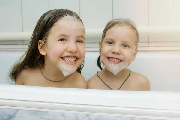 Två vackra små flickor i skum har roligt medan de sitter i badrummet. Lekfulla barn med skum på huvudet och ansiktet. Barnhygien. Badbarn tillsammans. Barnhygien. — Stockfoto