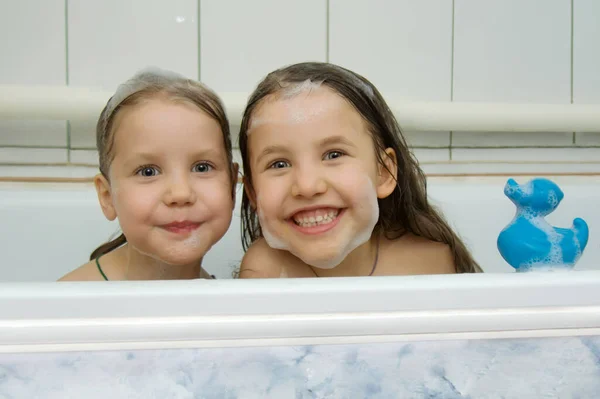 Två vackra små barn i skum har roligt medan de sitter i ett badkar med en badanka. Lekfulla tjejer med skum på huvudet och ansiktet. Barnhygien. Badbarn tillsammans. — Stockfoto