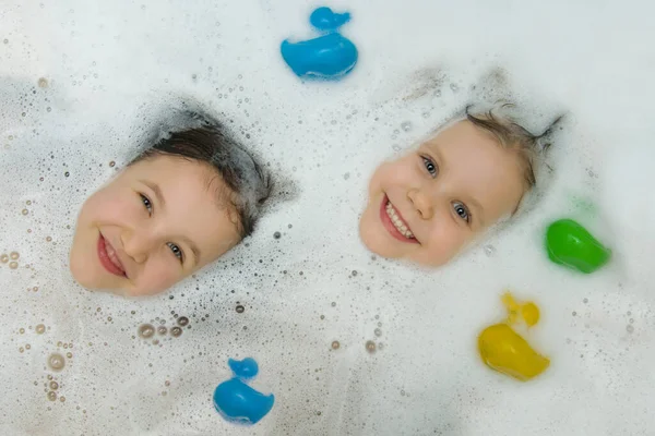 Las caras juguetonas de los niños asoman de la espuma, sonriendo alegremente mirando a la cámara. Dos chicas lindas se bañan juntas en un baño con patos para bañarse. Vista desde arriba. Higiene infantil. — Foto de Stock