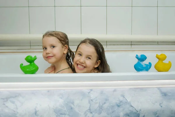 Två vackra små skumbarn har roligt när de sitter i ett bad med färgade ankor för bad. Badbarn tillsammans. Lekfulla flickor tittar upp ur badet och ler mot kameran.. — Stockfoto