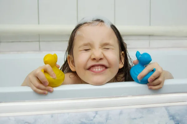 Barnhygien. En glad liten flicka med skum på huvudet tittar ut ur badkaret, blundar och håller ankorna för bad. — Stockfoto