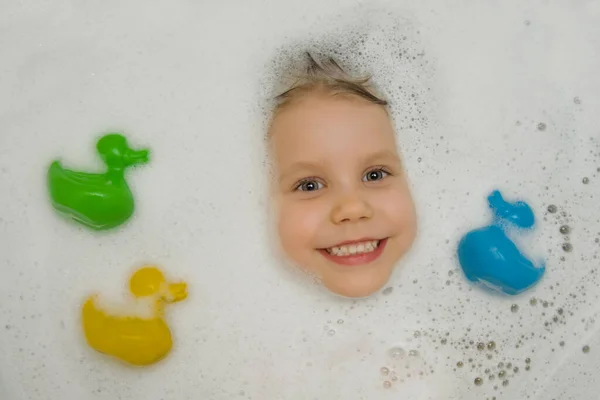 Egy vidám babaarc kukucskál ki a habból a fürdőszobában, és vidáman mosolyog a kamerába. A gyönyörű kislány fürdik sokszínű kacsákkal fürdéshez. Kilátás fentről. Gyermekhigiénia. — Stock Fotó
