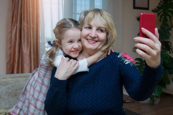 Großmutter und Enkelin umarmen sich und haben Spaß dabei, in ein Smartphone zu schauen. Eine erwachsene Frau von 50-60 Jahren macht ein gemeinsames Selfie-Foto mit ihrer kleinen schönen Enkelin. — Stockfoto