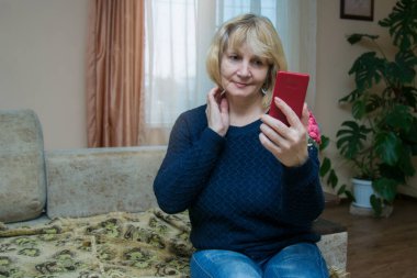 50-60 yaşlarında güzel bir kadın elinde telefonla akıllı bir telefona bakıyor ve evdeki kanepede oturuyor. Büyükannem bir video görüşmesi yapıyor. Emekliler için uygun fiyatlı modern teknolojiler.