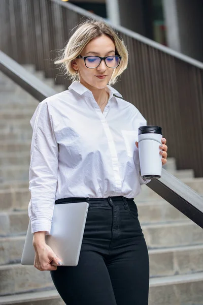 A business woman in a white shirt and black trousers descends the stairs. Young blond woman with coffee in a reusable cup, glasses and laptop.