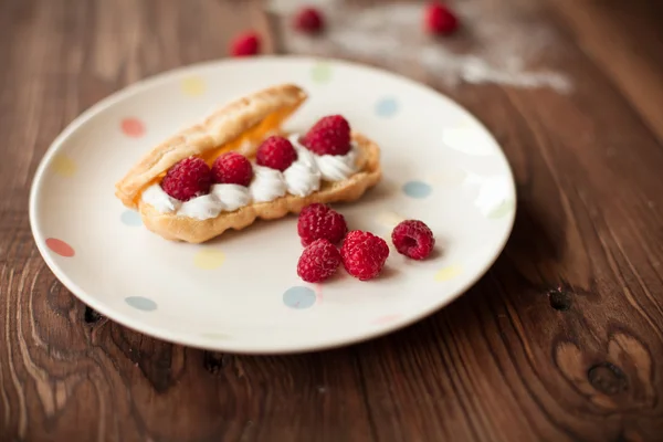 Cream eclairs with fresh raspberries on plate on wood table — Stock Photo, Image