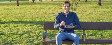 Mavi gömlekli genç adam not defterine bir bankta yazı yazıp gülüyor, arka planda günbatımı var. Parkta çalışan bir genç. Parkta mola verdim.