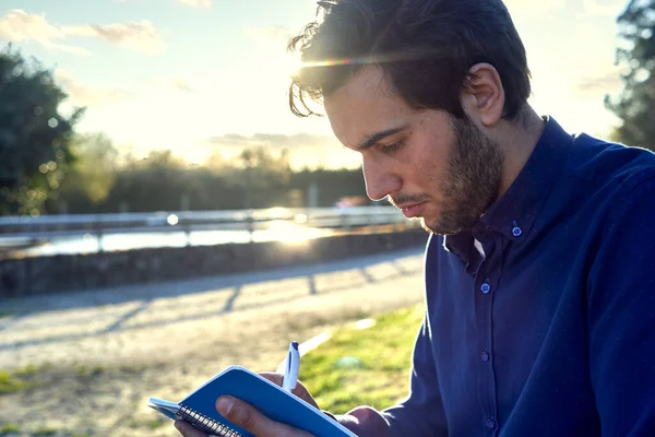 Mavi gömlekli genç adam not defterine bir bankta yazı yazıp gülüyor, arka planda günbatımı var. Parkta çalışan bir genç. Parkta mola verdim.