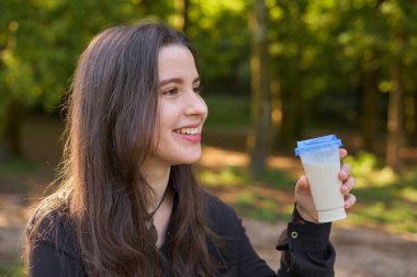 Uzun saçlı, güzel bir kız, doğada telefonunu taştan bir masa ve odaklanmamış arka planda kullanırken sütlü kahve içiyor. Sosyal medyasına bakarken parkta milkshake içen gençler.
