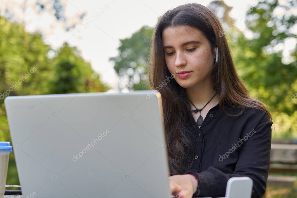 long-haired pretty businesswoman with laptop, headphones and coffee at stone table. teenager working on her laptop in nature with unfocused green background. girl with computer outdoors.