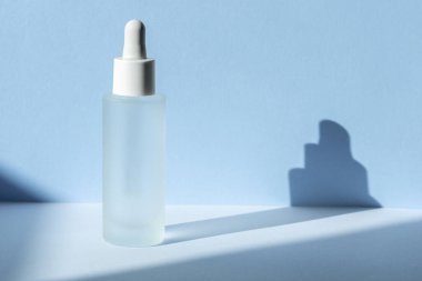 Cam şişedeki serum özü, mavi arka plandaki geometrik gölgeli hyaluronik asit sahnesi. Kozmetik ürün modeli. Cilt bakımı güzellik konsepti.