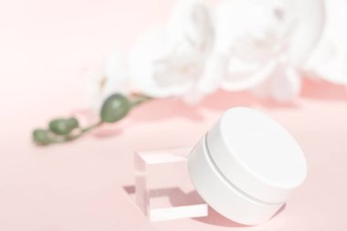 El kremi, nemlendirici, pembe çiçek arkaplanındaki beyaz kavanozda losyon, ön manzara. Yaşlanma karşıtı kozmetik ürün şablonu. Akrilik blok ve orkide dekoru