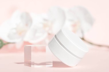 El kremi, nemlendirici, pembe çiçek arkaplanındaki beyaz kavanozda losyon, ön manzara. Akrilik blok ve orkide dekoru. Yaşlanma karşıtı kozmetik ürün şablonu, lüks vücut bakımı