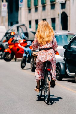 Milan, İtalya - 25 Mayıs 2024: Çiçekli elbise giymiş bir kadın güneşli bir günde şehirde bisiklet sürüyor. Araba ve motosikletli şehir trafiği. Yaz yaşam tarzı ve yavaş seyahat.