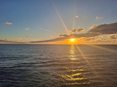 Sakin bir denizin üzerinde sakin bir günbatımı ufku aydınlatır. Güneş ışınları yumuşak bulutları keserken, suyun üzerinde sıcak altın bir parıltı oluşturur. Seyahat, doğa ve rahatlama temaları için ideal.