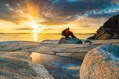 Şafak vakti kıyı koyundaki kayalıklarda duran sırt çantalı bir adamın görüntüsü.