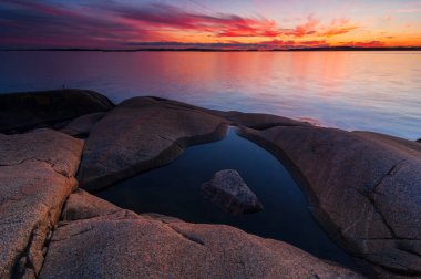 Bu güzel günbatımı, sakin sularda yansıyan renklerle dolu canlı bir gökyüzünü sergiliyor. İsveç 'in Göteborg şehrinde kıyı kayaları, gün geceye dönerken sakin bir ön plan oluşturur..