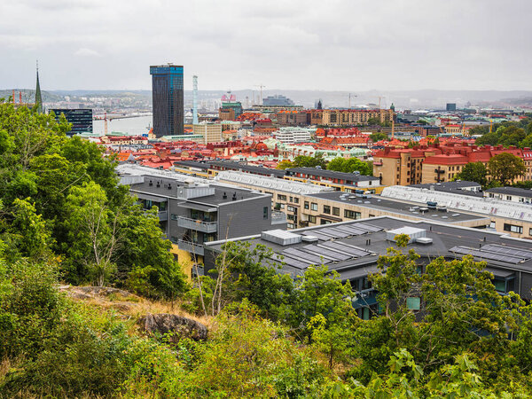 Пышная зеленая растительность обрамляет оживленный город Гётеборг, демонстрируя красные крыши и современные здания на фоне облачного неба. Вид запечатлевает гармонию между природой и городской жизнью.