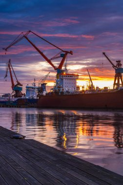 Göteborg 'da şafak vakti, vinçler kenetlenmiş gemilerin üzerinde dik dururlar. Canlı gün doğumu gökyüzünü turuncu ve mora boyarken su da güzel manzarayı yansıtıyor..