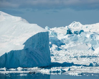 Sermeq Kujalleq 'teki büyük buzdağlarıyla çevrili Ilulissat fiyordundan bir tekne geçiyor. Sahne açık gökyüzünü ve yüzen buz parçalarını gösteriyor. Doğal manzarayı ön plana çıkarıyor..