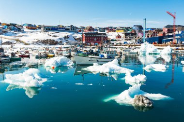 Ilulissat limanında büyük bir buzdağı yüzer. Yakınlarda küçük balıkçı tekneleri var. Sahilde renkli evler var. Su açık mavi gökyüzünü yansıtır. Ülkenin bazı kısımları karla kaplıdır..