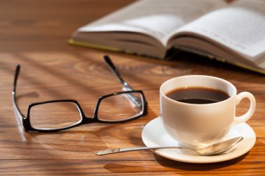 Fincan kahve, açık bir kitap ve bir masa üzerine gözlük.