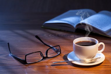 Fincan kahve, açık bir kitap ve bir masa üzerine gözlük.