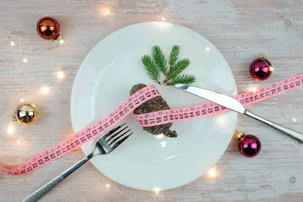 Il concetto di dieta natalizia. Coni di pino con metro a nastro su piastra bianca, forchetta e coltello su tavolo in legno, luci e palline, decorazione rami di pino. Idea menu vegetariano. Vista dall'alto. — Foto Stock