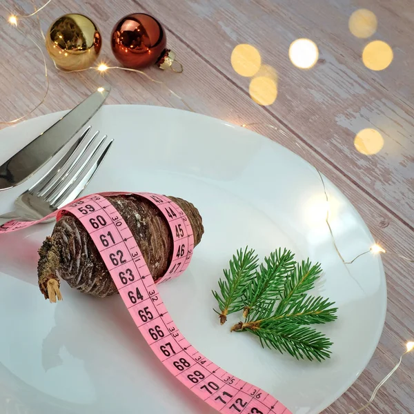 Christmas diet concept. Pine cones with tape measure on white plate, fork and knife on wooden table. Golden lights bokeh and balls decoration. Vegan menu concept. Square image.