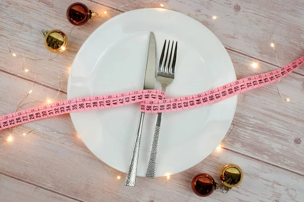 Noel yemeği için diyet konsepti. Şerit mezura, çatal ve bıçakla beyaz tabakta, tahta masada. Işıklar, bokeh ve toplar süsleme. Masa ayarı. Üst görünüm. — Stok fotoğraf
