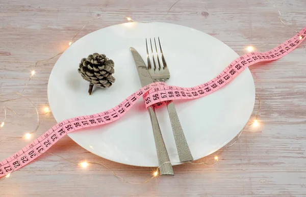 Dieta concetto per la cena di Natale. Il metro a nastro con forchetta e coltello su piastra bianca e pigna su tavolo in legno. Luci decorazione. — Foto Stock