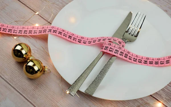 Dieta concetto per la cena di Natale. Il metro a nastro con forchetta e coltello su piastra bianca, su tavolo in legno. Luci, bokeh e decorazione palle. Impostazione della tabella. — Foto Stock