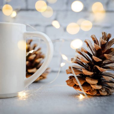 Beyaz bardaklar ve çam kozalakları rahat bir noel düzeninde. Tuğla beyaz duvar zemininde şenlikli ışıklar ve kare kare şeklinde altın bokeh.