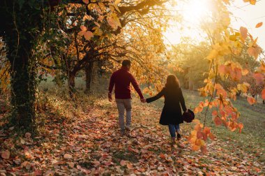 Sonbahar mevsiminde gün batımında bir parkta düşen yapraklarla el ele tutuşan çift..