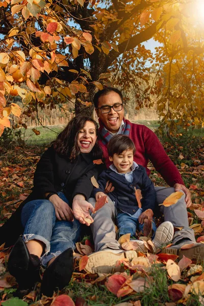 Multi ethnic playful family portrait sitting on fallen leaves in a park during autumn season. Happiness, togetherness concept.