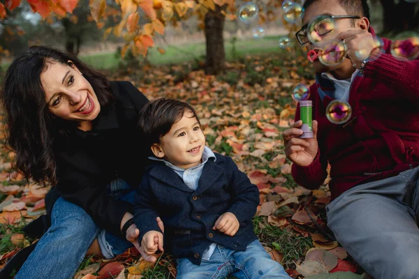 Multi-ethnic family portrait sitting in a park during Autumn season. Mother, father and son having fun doing bubbles soap and trying to break them. Togetherness, sharing moments, family time concept.