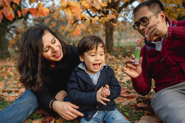 Multi-ethnic family portrait sitting in a park during Autumn season. Mother, father and son having fun doing bubbles soap and trying to break them. Togetherness, sharing moments, family time concept.