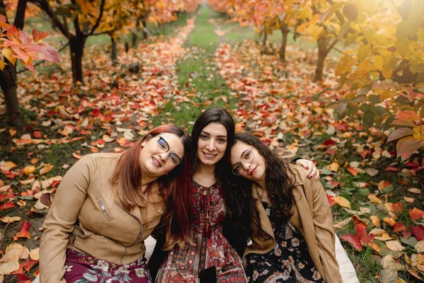 Three girls smiling portrait sitting on the ground in a field during fall season. Togetherness, Autumn season, friendship, red and orange color concept.