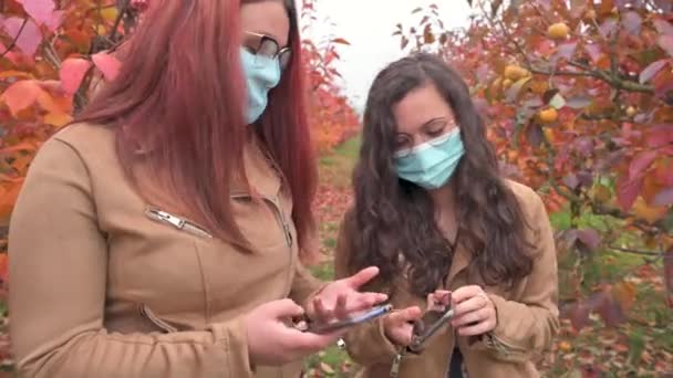 紅葉の季節には2人の女の子がスマホを使って笑顔と会話をしています コロナウイルスパンデミック時の保護面 — ストック動画