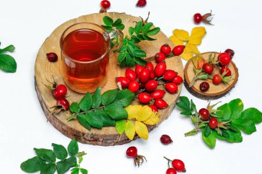 Tahta testerenin üzerinde, beyaz bir yüzey üzerinde kuru ve taze gül üzümlerinin yanında, cam bir fincanda sağlıklı bir gül çayı.