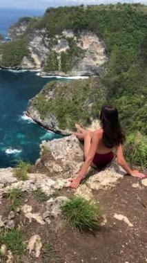 Bali adasında, okyanusun yukarısındaki uçurumun kenarında oturan mor mayolu kadın.