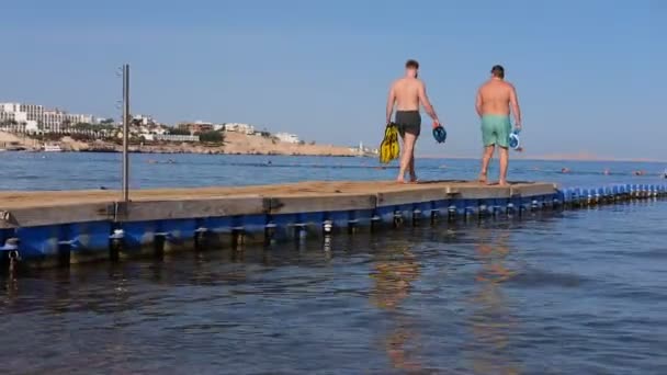 Чоловіки в плавальних шортах ходять по дерев'яному і пластиковому пірсу вздовж моря. . — стокове відео