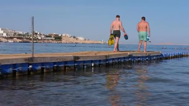 Чоловіки в плавальних шортах ходять по дерев'яному і пластиковому пірсу вздовж моря. . — стокове відео