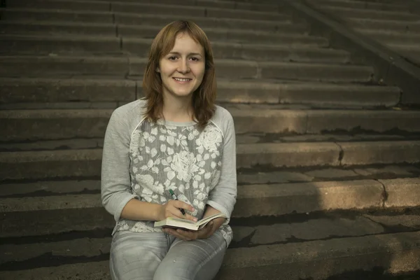 Foto de chica bonita sentada en las escaleras de la universidad y escribiendo algo —  Fotos de Stock