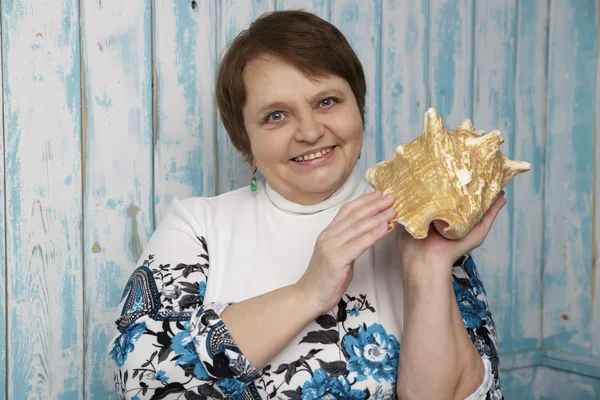 Una anciana sosteniendo una concha. Enfoque selectivo en su cara. Fondo marino azul. Mujer de sesenta años . — Foto de Stock