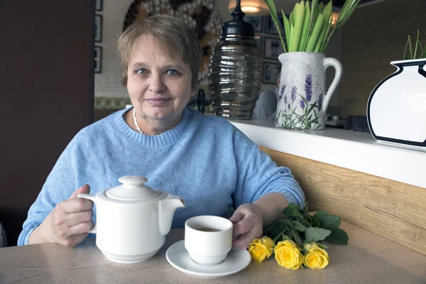 Gammal kvinna klädd i en blå tröja som sitter i kaféet med vattenkokare och — Stockfoto