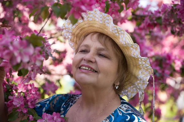 Smiling adult woman wearing a hat under pink flowering tree in garden — Stock Photo, Image