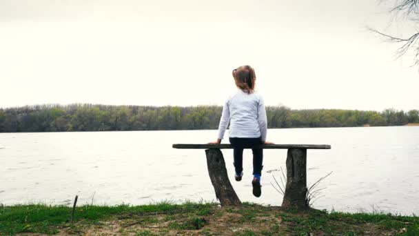 Linda niña se sienta en el banco cerca del río — Vídeos de Stock